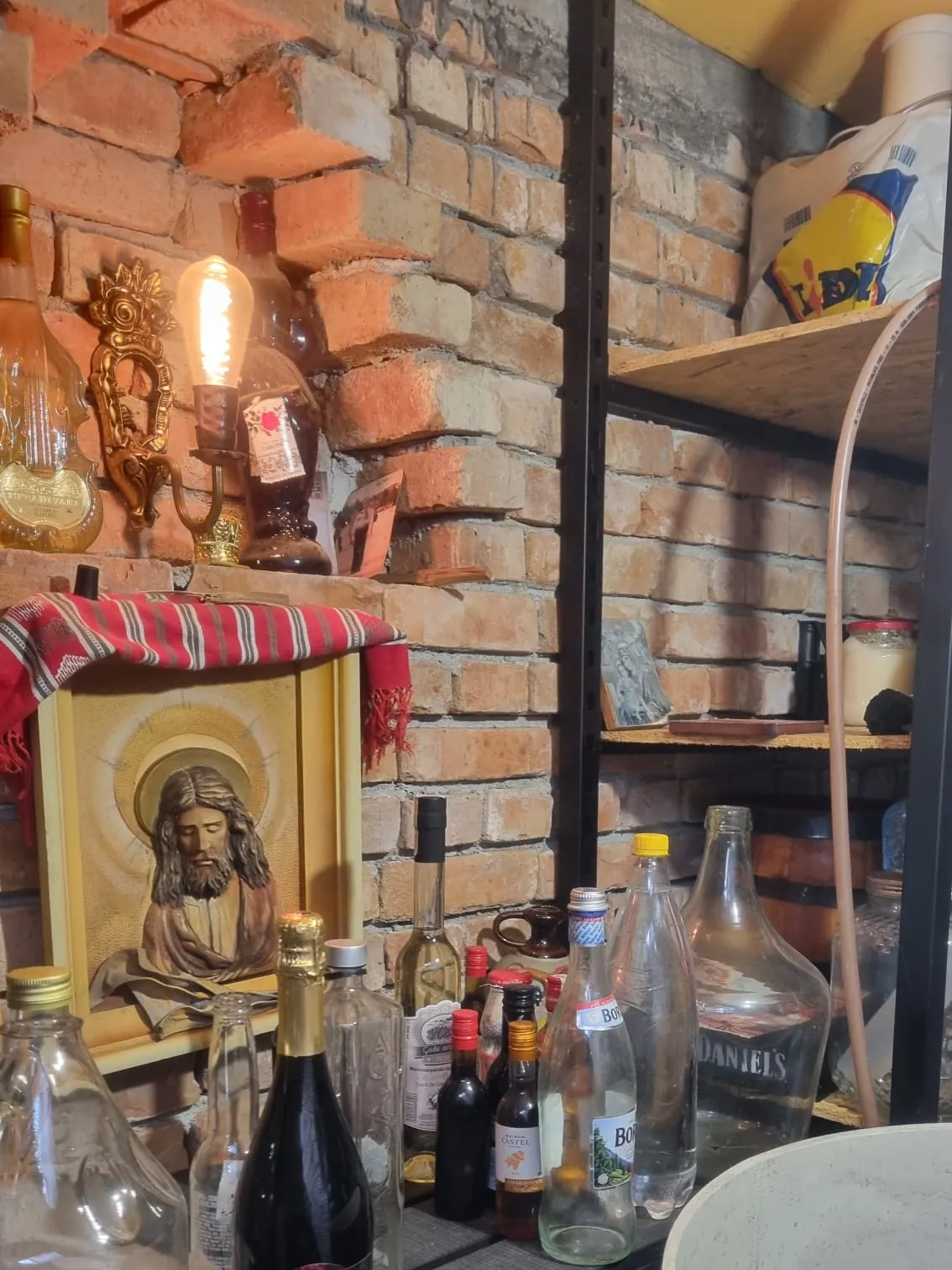 Interiorul unui beci terminat, cu pereți de cărămidă, lampă caldă și rafturi cu damigene și sticle
