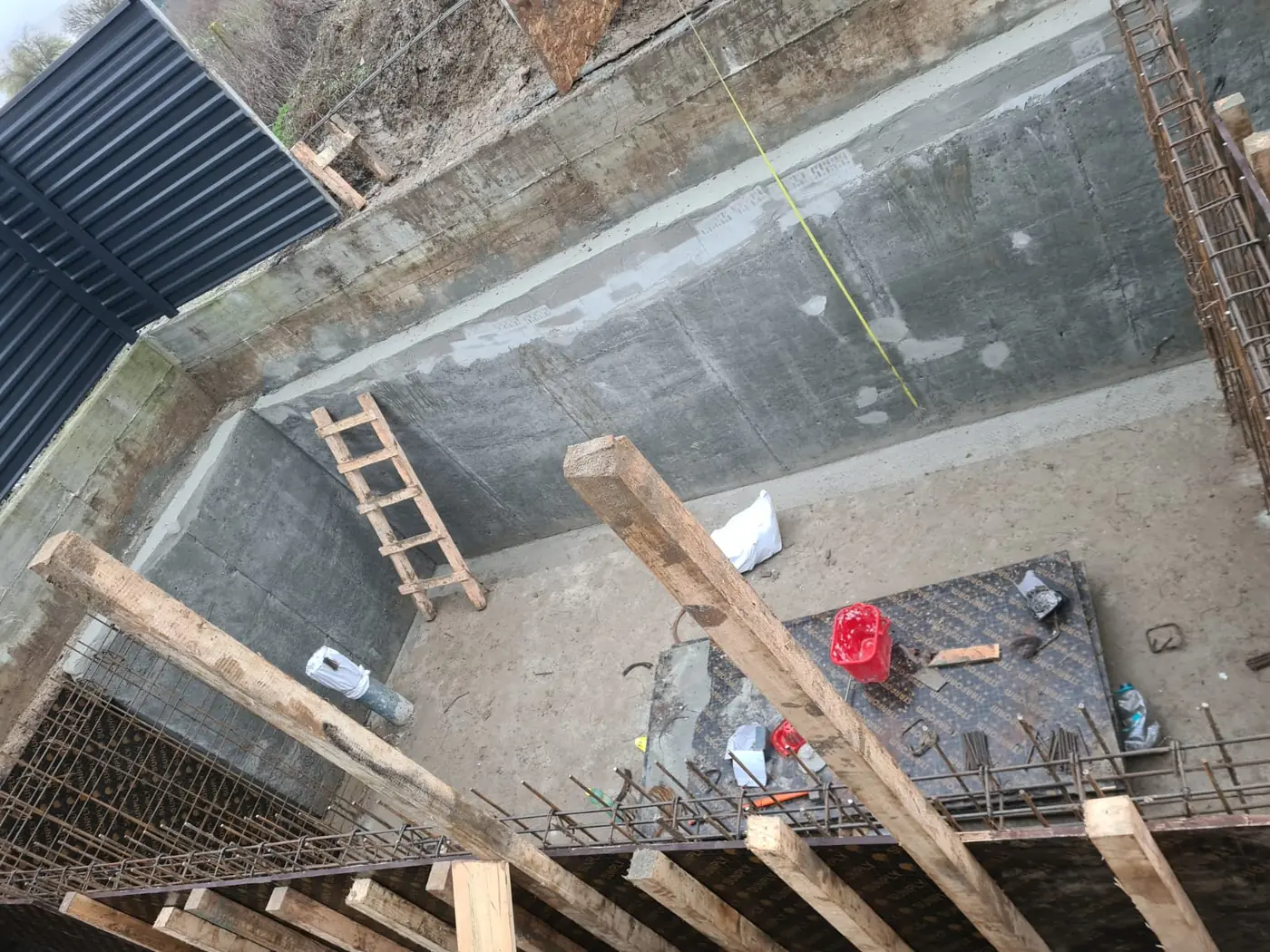 Pereți de beton turnați ai unui beci, cu scara de acces sprijinită de perete și armătura pregătită pentru placă